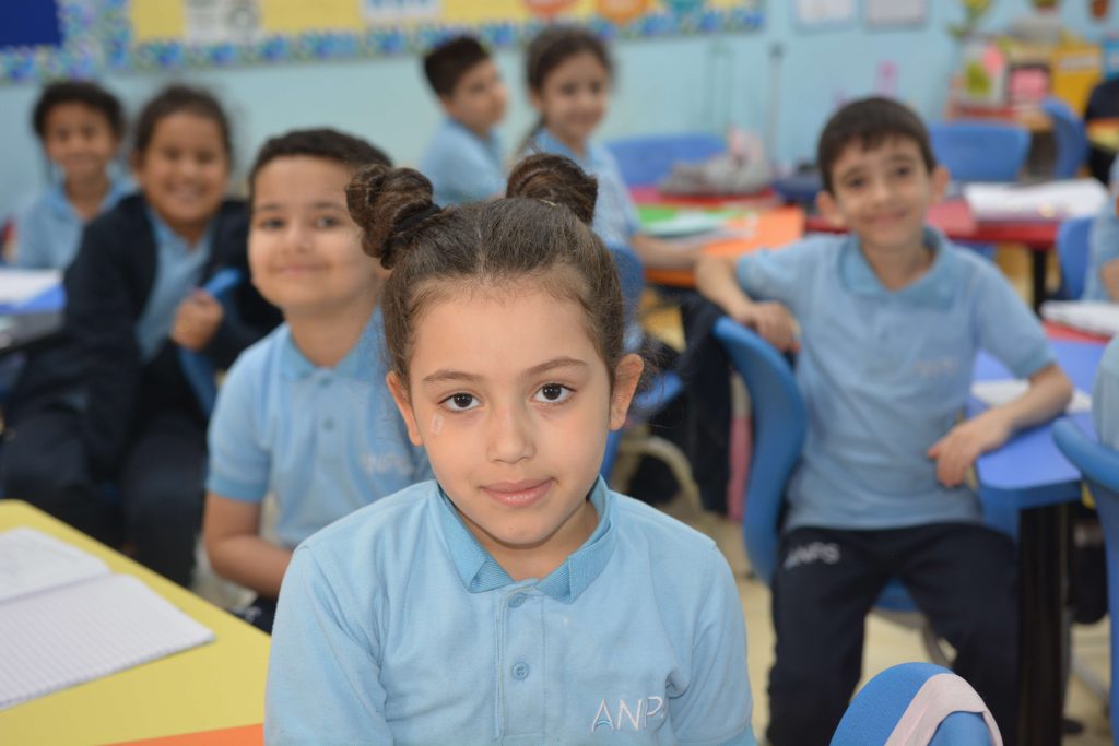 An ANPS Primary pupil at her desk looking towards the camera while classmates work behind her