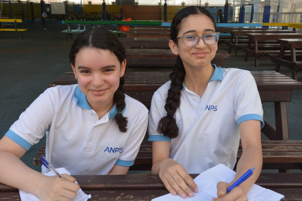 Two ANPS Middle School students in white school polo shirts writing at an outdoor bench