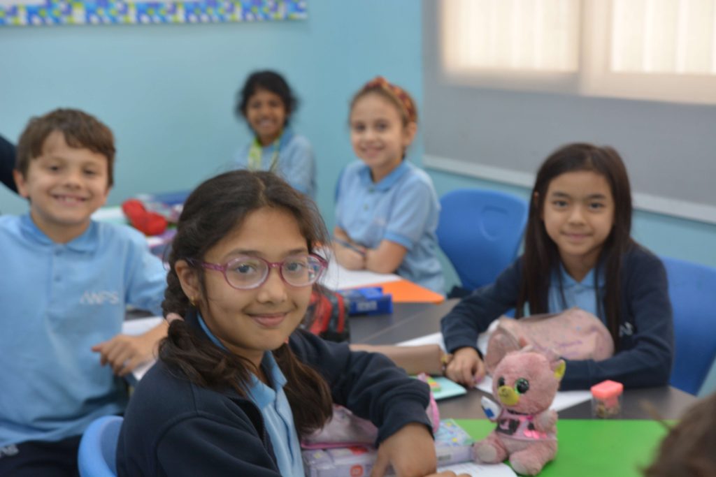 ANPS Primary students in light blue polo shirts and navy cardigans smiling at their classroom desks