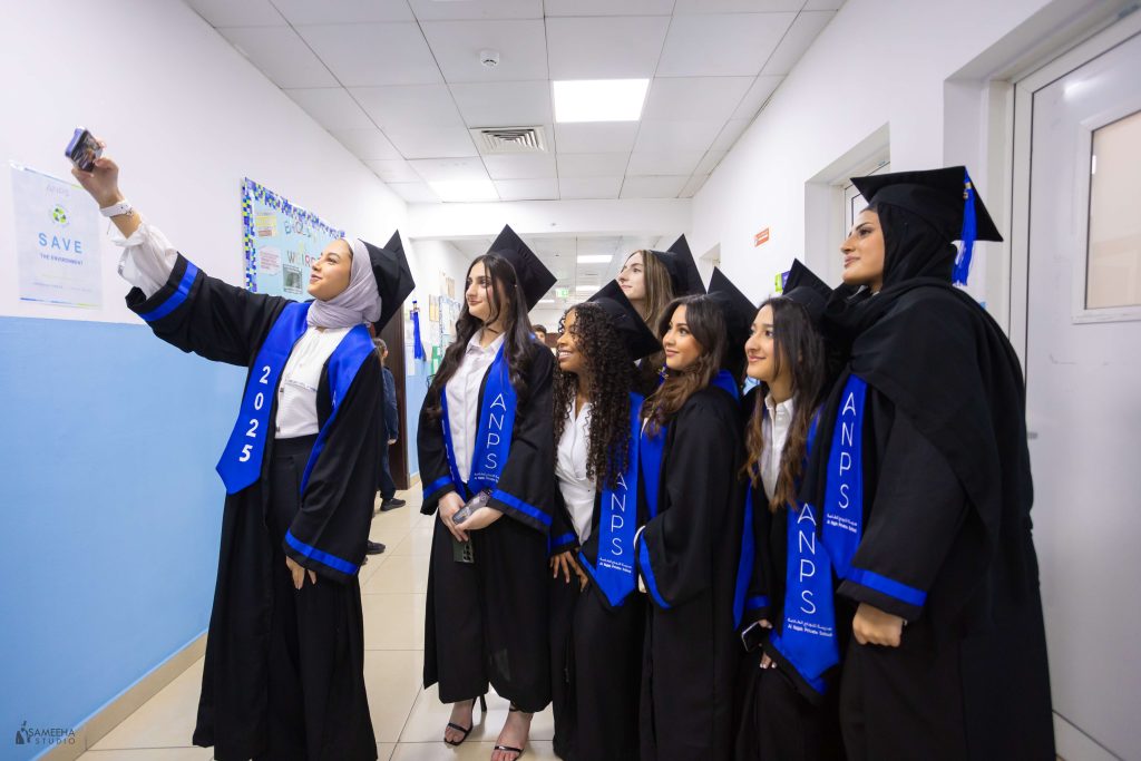 ANPS graduates in black gowns and blue 2025 sashes taking a group selfie in a school corridor